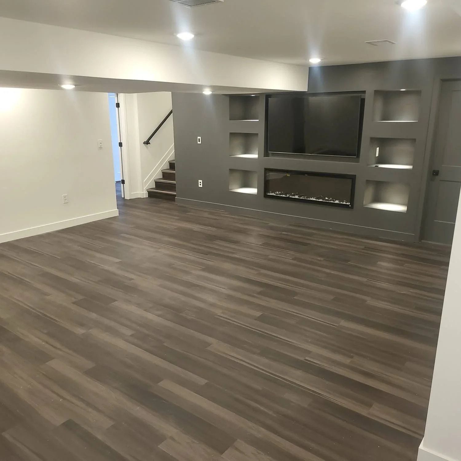 Finished basement with dark wood flooring, gray drywall entertainment wall, built-in shelving, electric fireplace, and recessed lighting