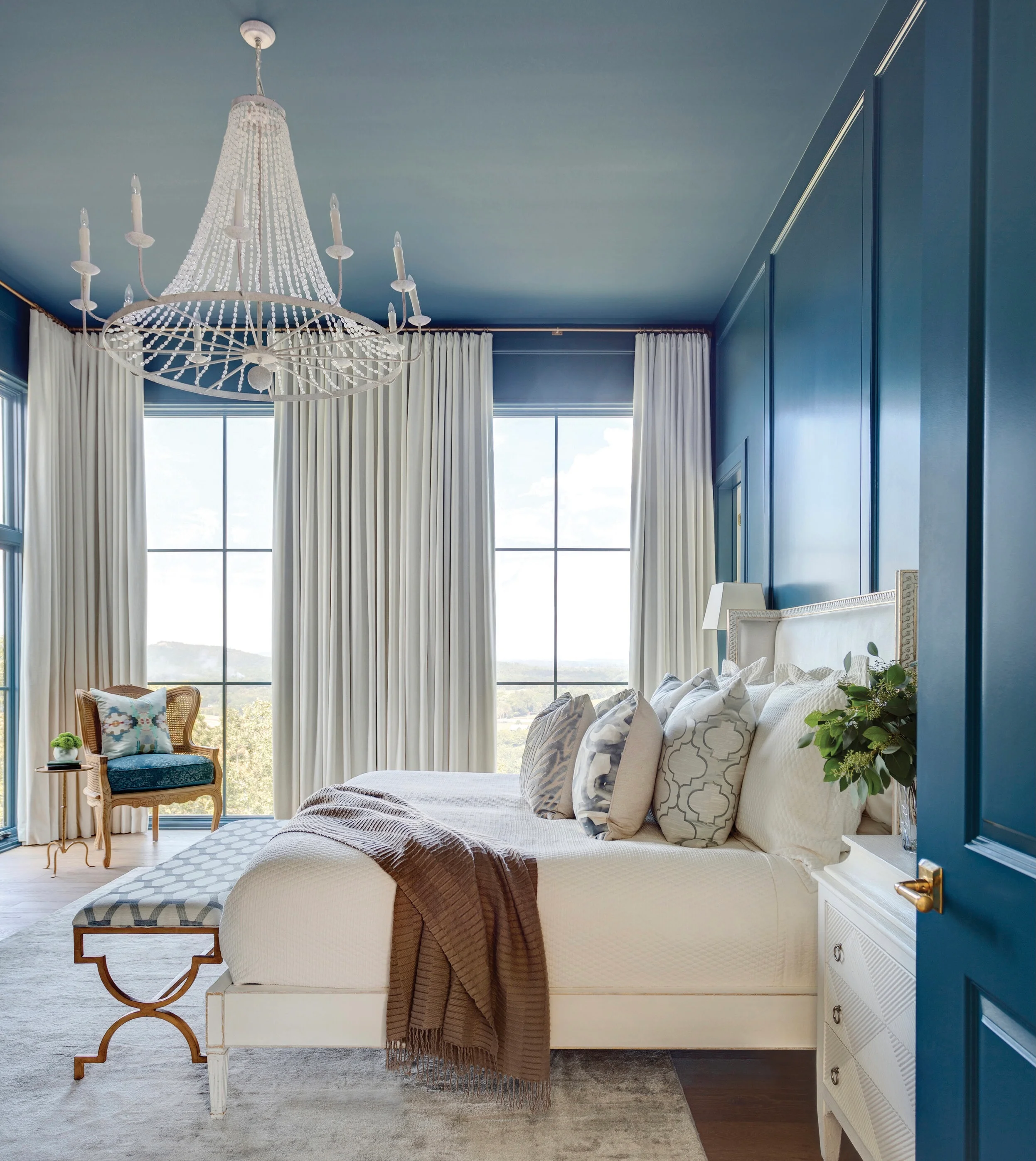 Master bedroom with blue walls, beaded chandelier, cream curtains, and white upholstered bed