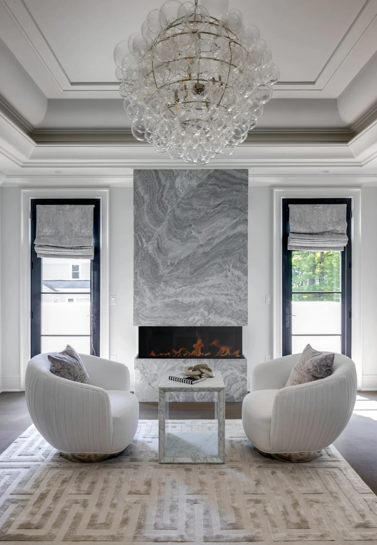 Modern living room with marble fireplace, glass chandelier, and white curved chairs